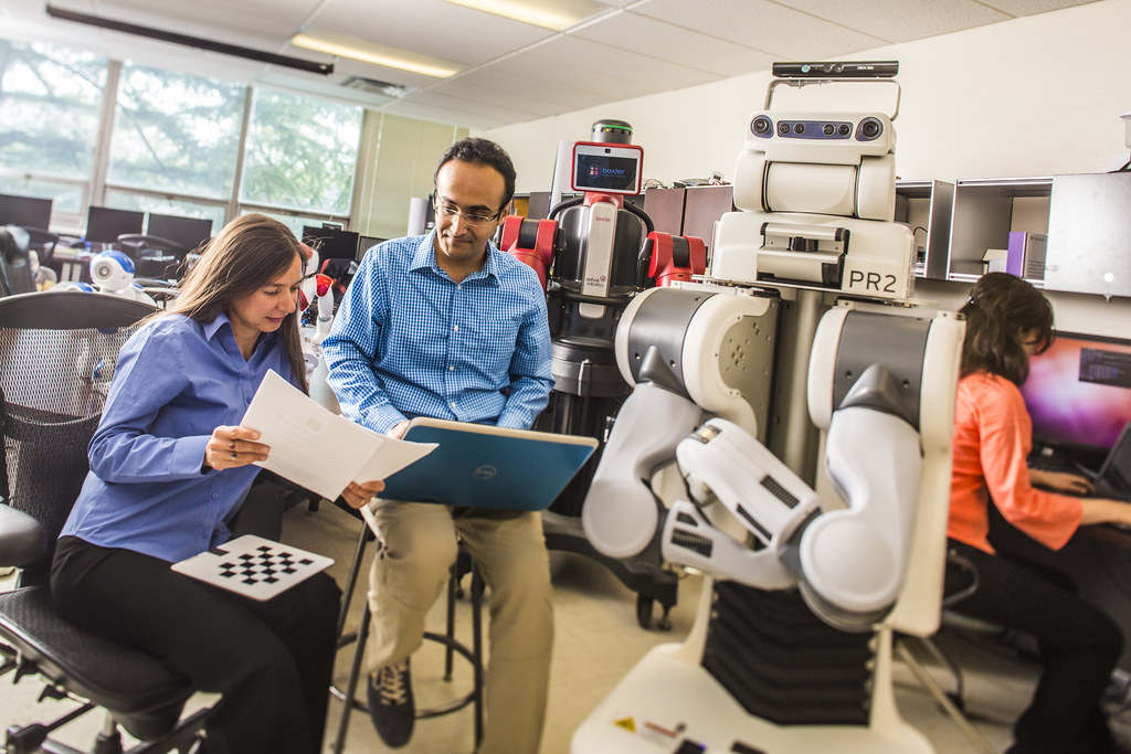 Two professionals discussing reports in the ENG Robotics room next to a PR2 robot. One professional is taking notes on a laptop.