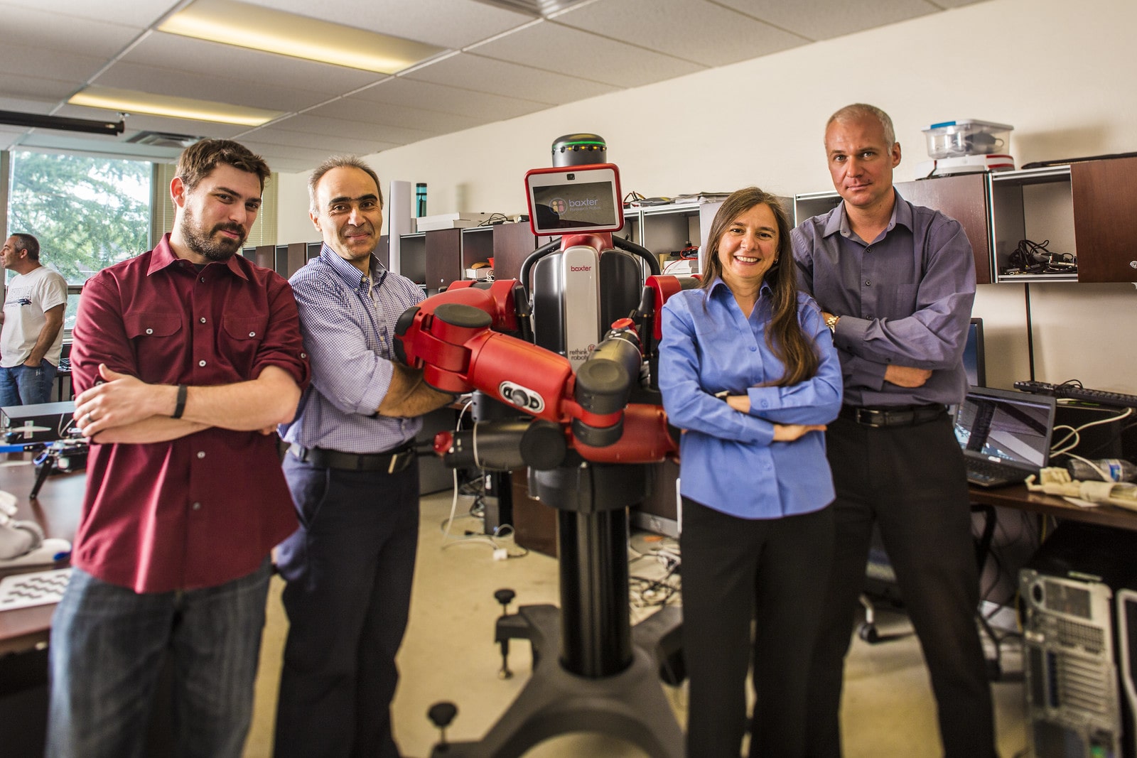 Engineers and other professionals standing inline with a red Baxter robot in a well lit robotics lab.