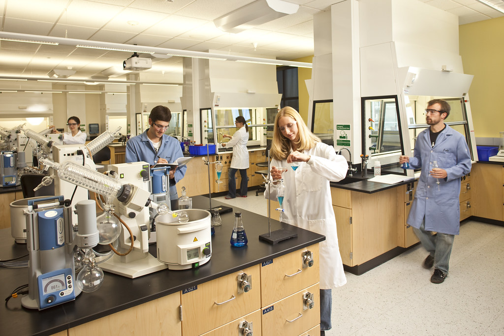 Multiple students in a laboratory. two students are holding and mixing blue liquid solution in beakers. The others are taking notes.