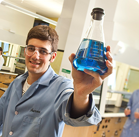 Multiple students in a laboratory mixing beakers with solutions of various mixtures. A student in the foreground is holding a large beaker containing a blue solution.